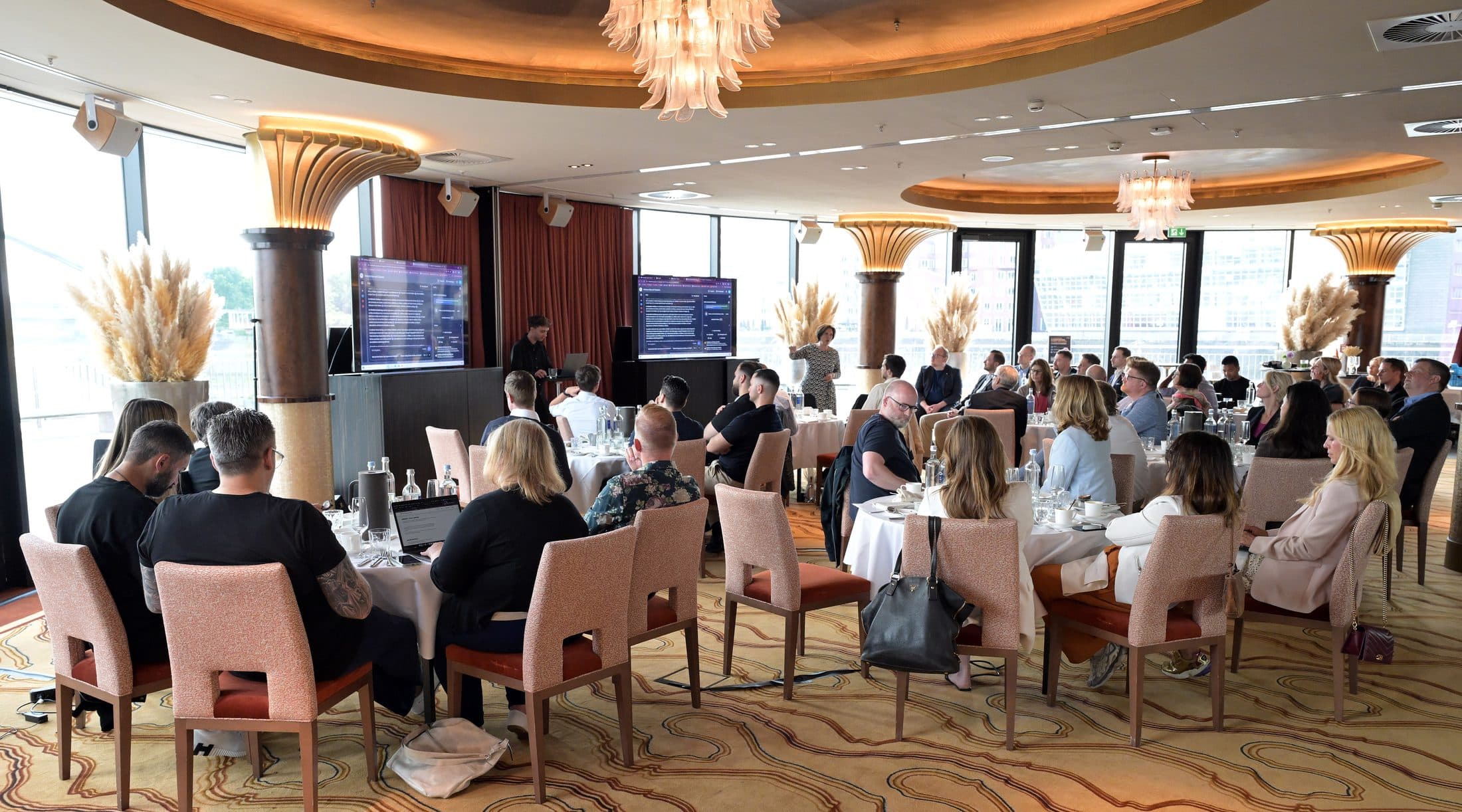 Voll besetzter Saal im Hyatt Düsseldorf, vorne läuft eine KI-Session auf zwei Bildschirmen
